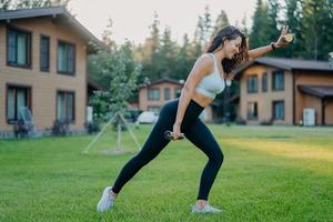 Photo of active slim woman does sport exercises with dumbbells, dressed in cropped top, leggings, sneakers, leads active lifestye, poses outdoor near houses on green grass being in good physical shape
