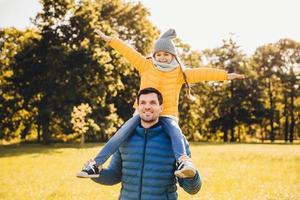 Handsome father wears anorack gives piggyback to his little funny girl, stroll together outdoor, enjoy sunshine. Attractive dad and his daughter who rides on his back, gestures, feels freedom photo
