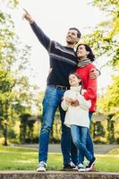 Vertical portrait of family of three members stand close to each other, embrace, have walk across autumn park, notice squirrels on tree. Friendly father, mother and daughter have good relationship photo