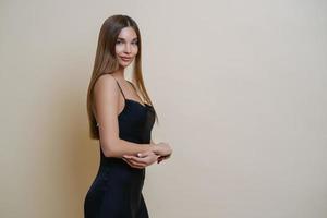 Beautiful slim woman stands in profile, wears elegant black dress, has long straight hair, looks with charming expression at camera, poses against beige background, copy space for your information photo