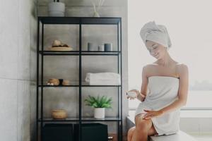 Photo of relaxed beautiful young woman applies body cream at home, uses cosmetic product for healthy skin, poses in bathroom, wearrs wrapped towel on head and over body. Beauty treatment concept