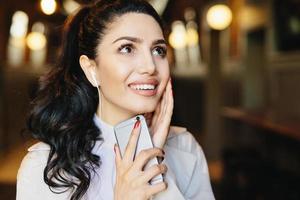 Fashion lifestyle concept. Portrait of cute woman with pony tail having charming smile listening to music with earphones using mobile phone while resting in cafe. Caucasian brunette female with mobile photo