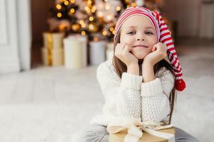 Horizontal portrait of adorable little child, leans on hands with present box, sits against decorated Christmas tree. Blue eyed small kid in knitted white sweater poses at camera. Holidays concept photo