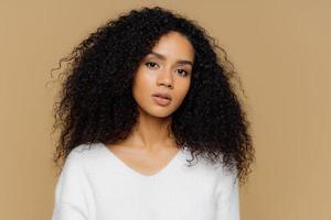 Portrait of serious beautiful dark skinned female with frizzy black hair, has minimal makeup, looks calmly at camera, wears white jumper, stands against brown background, being deep in thoughts. photo