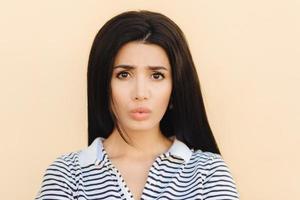 Gloomy brunette young female pouts lips in discontent, being dissatisfied with something, has healthy pure skin, dark eyes and make up, dressed in casual clothes, isolated over studio background photo