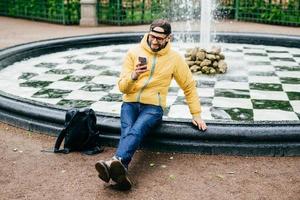 Leisure and free time concept. Portrait of carefree man with beard dressed in casual clothes leaving his rucksack near fountain using smartphone checking his e-mails having walk in beautiful park photo