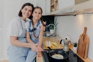 Lovely mother and daughter in aprons embrace and smile happily, fry eggs on modern stove in kitchen, use frying pan, prepare tasty breakfast. Family, children, motherhood and cooking concept photo