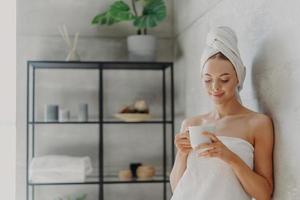 Fresh young European woman wrapped in towel after bath, stands in bathroom with mug of coffee or tea, enjoys morning, has pleased face expression, healthy glowing skin. Hygiene routine concept photo