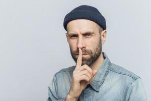 Serious pleasant looking bearded male with mysterious expression keeps fore finger on lips, asks to keep personal information confidential, wears stylish hat and denim shirt, isolated on grey wall photo