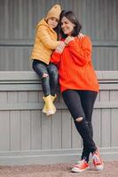 Vertical shot of pretty beautiful young woman dressed in red sweater and ragged jeans, stands closely to her small daughter who embraces her, enjoy togetherness. Parenthood and childhood concept photo