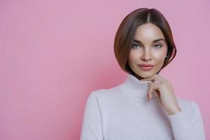 Portrait of brunette woman keeps hand on collar of turtleneck, has healthy perfect skin, wears minimal makeup, has serious expression, poses against pink background. Human facial expressions concept photo