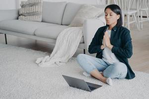 Young woman practicing yoga online at home. Girl sitting at laptop in lotus pose and meditating. photo