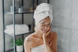 Beautiful European woman with makeup touches skin, has minimal makeup, has healthy glowing skin, wrapped in bath towel, enjoys rest at home. Spa woman poses in bathroom. Beauty, wellness concept photo