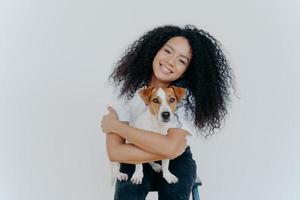 Horizontal shot of pleasant looking curly young woman embraces lovely pet, holds cute dog, smiles broadly, tilts head, isolated against white background. People, animals, good emotions concept photo