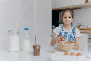 Adorable European child enjoys cooking activity, breaks egg in bowl, whisks ingredients, prepares tasty pastry, being young cook, wears apron, poses against kitchen interior, learns how to make cake photo
