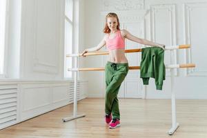 foto completa de una modelo femenina en forma con el estómago desnudo, se inclina en la barra de ballet, se calienta en el estudio de baile, se viste con ropa deportiva y zapatillas rosas, tiene buenas habilidades. gente, hobby, concepto de gimnasia