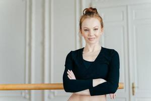 tiro de cintura arriba de una bailarina de ballet segura de sí misma y satisfecha que cruza las manos sobre el pecho, viste una camiseta negra, tiene el pelo pelirrojo, se para cerca de las barras, posa sola en el gimnasio, usa una máquina coreográfica foto