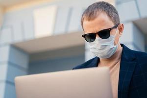 Close up shot of adult man wears sunglasses and medical mask, reads news online on laptop computer, finds out symptoms of coronavirus, has online communication during quarantine and self isolation photo