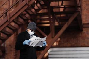 el hombre concentrado usa gafas de sol y abrigo, máscara médica, guantes de goma protectores, lee noticias recientes sobre la propagación del coronavirus del periódico, posa contra un fondo borroso con escaleras. foto