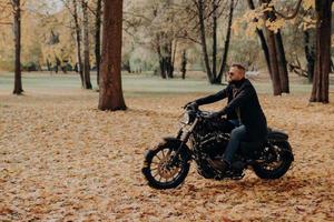 Confident male biker sits on motorbike, enjoys traveling on own transport, wears protective sunglasses, comfortable clothes, rides through trees in autumn park. Fall season, hobby, lifestyle photo
