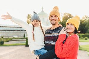 Cute lovely girl being on fathers hands, wave her hand as sees something wonderful into distance, spends her free time with father and mother, pose outdoors. Family embrace each other with great love photo