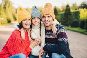 Outdoor portait of beautiful brunette female, handsome man, pretty small girl embrace together, have pleasant mood, walk in park, enjoy calm atmosphere and splendid sunny weather. Friendly family photo