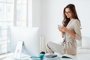 Elegant woman in beige formal suit reads news on website attentively, sits at desktop alone in cabinet, computer monitor and notepads around. Female employee uses modern smartphone in office photo