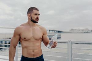 Muscular build bearded man goes in for sport, holds bottle of fresh water, focused into distance, poses on bridge near river during cloudy day, takes break after physical exercises. Cardio workout photo
