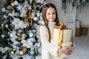 Indoor shot of adorable cute pretty small female child wears warm knitted clothes, holds huge wrapped present gift, recieves from Santa Claus, stands near decorated beautiful New Year tree