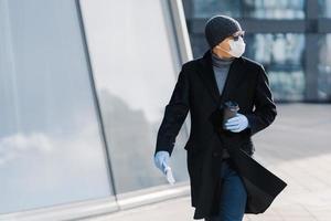 Shot of young man poses outdoor at street, looks aside, wears sunglasses, rubber gloves, medical mask during coronavirus outbreak, drinks hot beverage, tries to avoid public places during epidemic photo