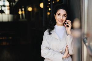 Elegant brunette businesswoman with dark eyes and full lips dressed in white clothes communicating over telephone having charming smile looking up with thoughtful expression. Technology concept photo