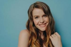 Studio portrait of cute teen girl with wavy hair smiling and looking at camera, isolated on blue photo