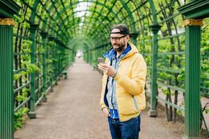 Trendy guy with beard and moustache wearing anorak, jeans and cap standing sideways having attentive look into his smartphone isolated over green arch. Human, style and modern technology concept photo