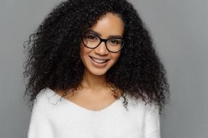 Headshot of curly haired smiling woman has healthy dark skin, Afro hairstyle, smiles gently at camera, weras optical glasses and white jumper enjoys spare time with friends isolated on grey background photo