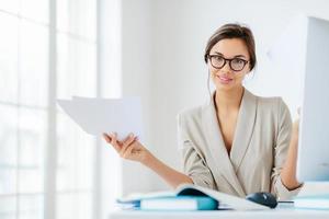 Indoor shot of good looking female in formal suit, holds paper documents, works on organization and creating startup project, poses at desktop with computer, checks information for presentation photo