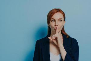 Young mysterious redhead europian female asking to keep silence and looking aside, isolated on blue photo