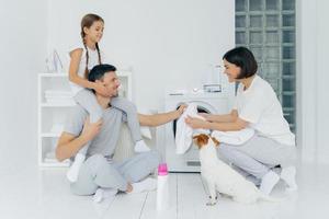 el padre feliz le da la espalda a la hija, ayuda a la esposa a cargar la lavadora, su mascota posa cerca, una botella de detergente en el piso blanco. familia amistosa en la lavandería, lavar la ropa, hacer las tareas del hogar foto