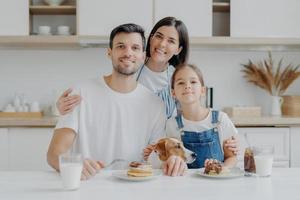 familia, ocio, pasatiempo, concepto de alimentación. padre, madre e hija, el perro jack russell terrier posan todos juntos frente a la cámara contra el interior de la cocina, disfrutan comiendo panqueques bebiendo leche fresca foto