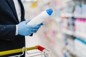 Man chooses detergent in household goods store, holds bottle with liquid powder, wears medical gloves to protect from coronavirus, reads product information. Making shopping during quarantine photo