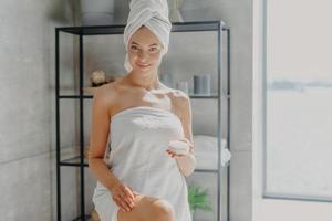 Horizontal shot of pleased smiling woman wrapped in bath towel, touches legs, has silky skin, uses cosmetic product after taking shower, has epilation procedure. Female applies moisturising cream photo