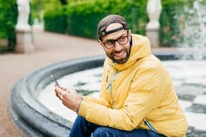 Sideways outdoor portrait of cheerful bearded stylish man in yellow jacket and eyeglasses sitting near fountain in park holding modern decive in hands looking happily aside admiring beauty of nature. photo