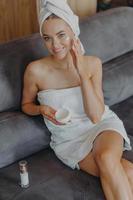Vertical shot from above of good looking cheerful European woman applies face cream on cheek, uses cosmetic lotion, smiles gently, wrapped in white bath towel, poses on comfortable sofa at home photo