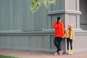 Horizontal view of fashionable European female in red loose sweater, ripped jeans, holds hands together with her younger sister, pose outside together, have walk in open air, have friendly relations photo