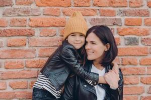 Horizontal shot of charming friendly little child and her mother embrace each other, dressed in black leather jackets, have appealing look and pleasant smile pose over brick wall. Relationship concept photo