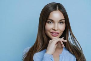 Isolated shot of attractive brunette woman with calm expression, healthy skin and makeup, keeps hand under chin, cares about complexion, looks beautiful, isolated on blue background, models in studio photo