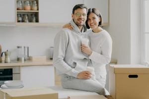 Image of glad family couple hug and stand closely to each other, drink takeout coffee, look with smile at camera, dressed in casual clothes, surrounded with cardboard boxes, spend free time in kitchen photo