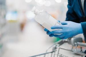 Unrecognizable person in medical gloves holds bottle of sanitizer gel, poses in shop, leans at shopping cart, buys product for coronavirus protection, blurred background. Disinfection, prevention photo