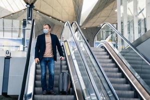 Man wears medical face mask, poses on escalator in airport, arrives from abroad, holds mobile phone and suitcase, protects from pandemic disease. Threat of epidemic in 2020, dangerous traveling