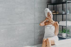 Relaxed young Caucasian female model wears towel wrapped on head, feels refreshed after taking shower, has healthy clean soft skin, poses in cozy bathroom. Women, beauty and hygiene concept. photo