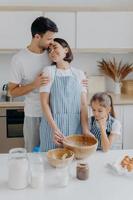 Happy lovely family in home kitchen, father embraces mother with love, little girl looks in bowl, observes how mommy cooks and whisks ingredients, use eggs for making dough. Domestic atmosphere photo